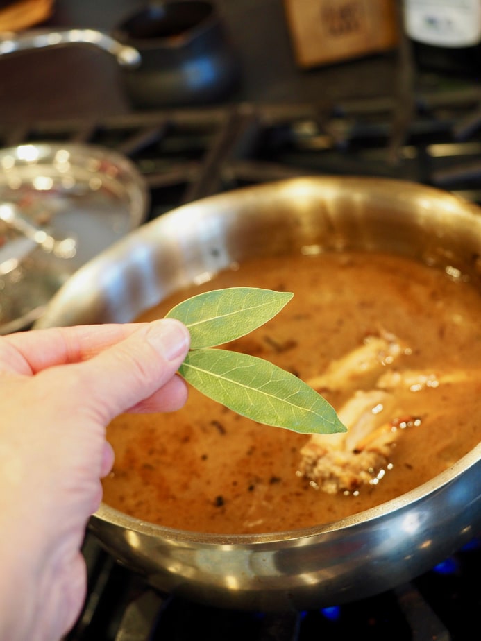 Hand holding two fresh bay leaves with skillet in background with giblets and gravy for turkey gravy stock. 