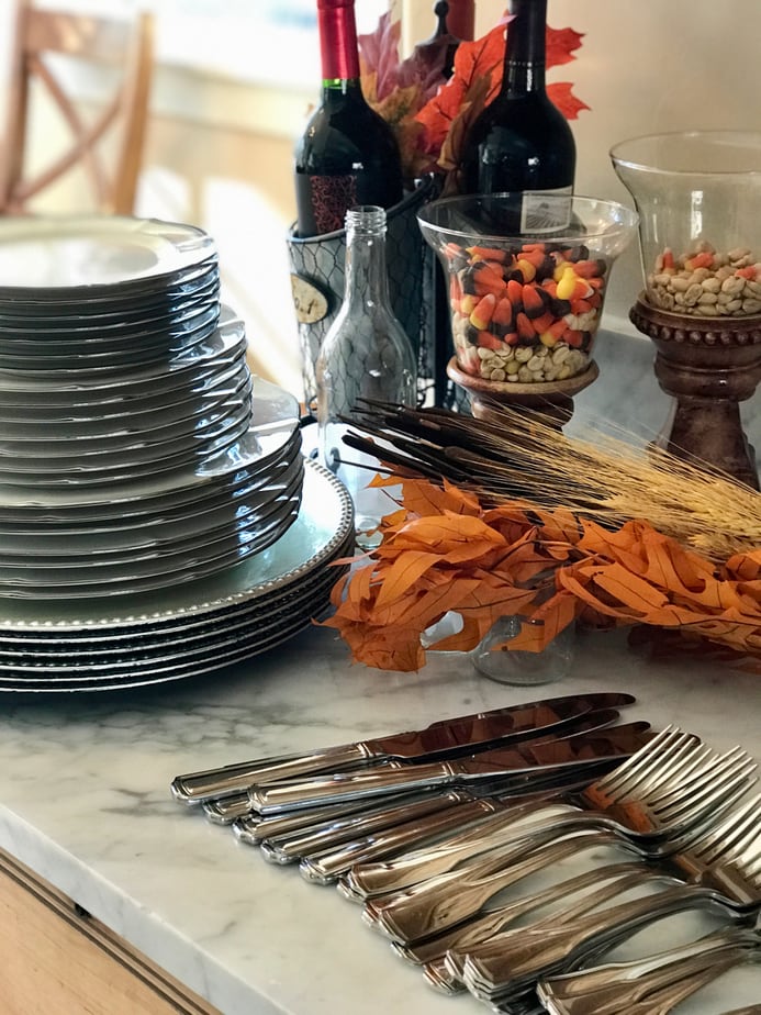 Set out everything needed for setting the table nearby. Chargers, stacks of plates, silverware with fall foliage in background.