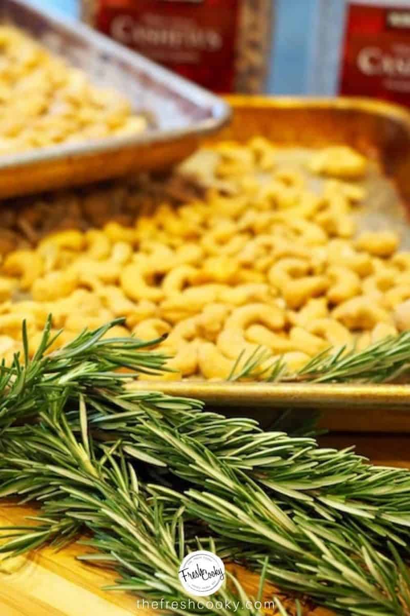 two sheet pans of cashews, with several stems of fresh rosemary herb sitting on a cutting board.