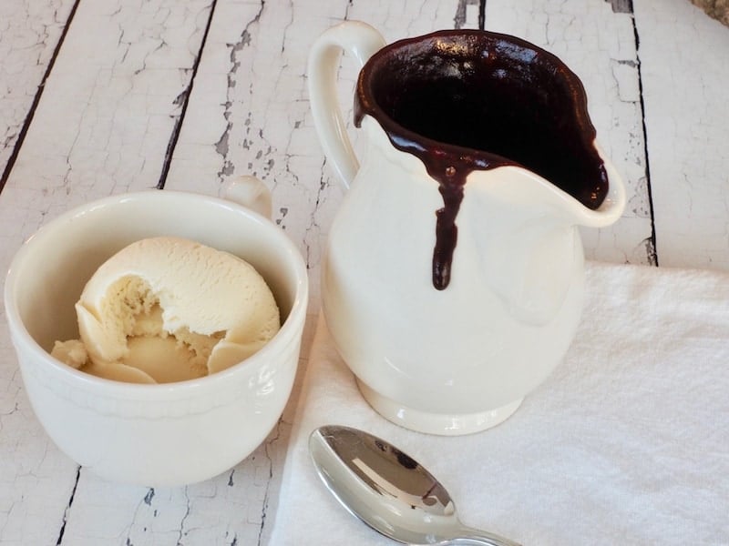 Chewy hot fudge in pitcher with bowl of vanilla ice cream and spoon next to it, on wooden board. Chewy Hot Fudge Sauce | thefreshcooky.com