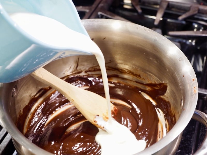 Pouring cream from a turquoise liquid measure cup into melted chocolate in small sauce pan with wooden spoon, old-fashioned hot fudge sauce.