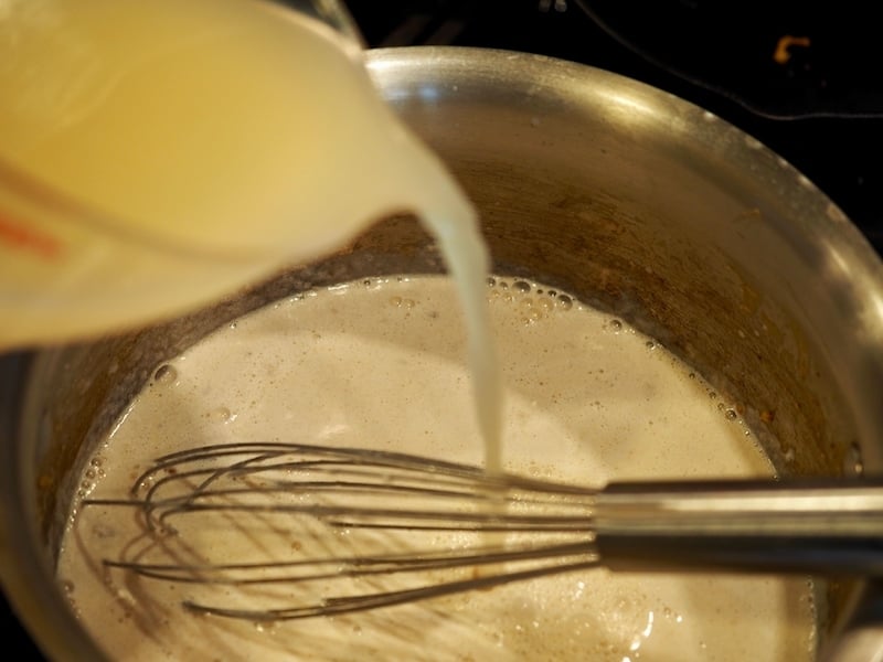 Pouring chicken stock into sauce pan filled with thickened roux and whisk sitting on side 