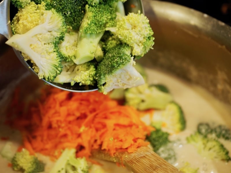 A spoon scooping in broccoli florets on top of shredded carrots in pot with wooden spoon 
