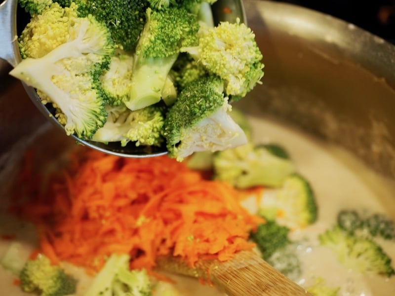A spoon scooping in broccoli florets on top of shredded carrots in pot with wooden spoon