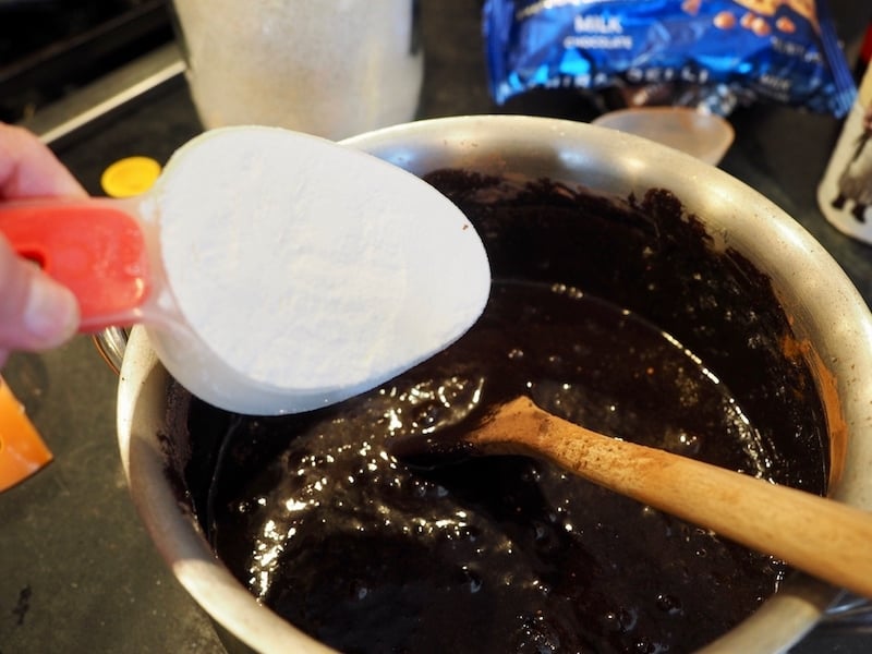 flour in measuring cup, over a pot with chocolate brownie mixture and a wooden spoon in the pot.