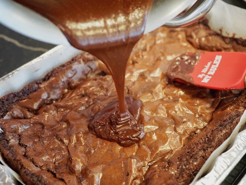 pouring hot frosting over the top of a pan of brownies with red spatula in the background.