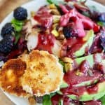white plate on sliced wood charger with a bright salad with greens, avocados, chicken, blackberries and a luscious blackberry vinaigrette along with two discs of fried goat cheese