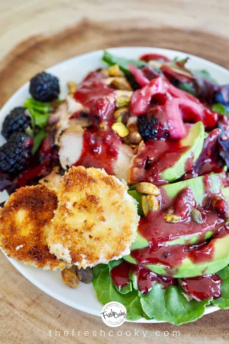 white plate on sliced wood charger with a bright salad with greens, avocados, chicken, blackberries and a luscious blackberry vinaigrette along with two discs of fried goat cheese