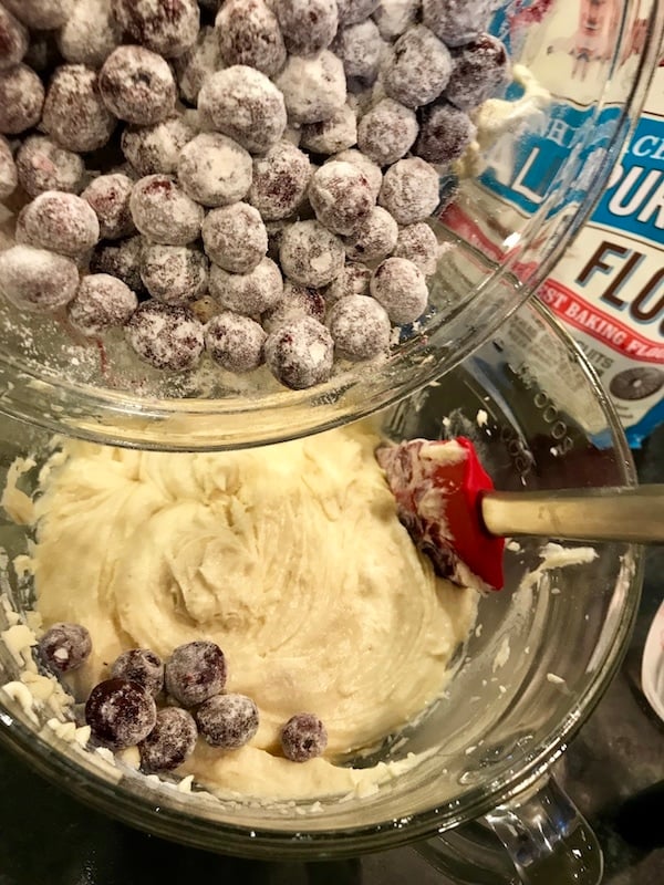 Glass mixing bowl with red spatula folding blueberries into blueberry cake dough.