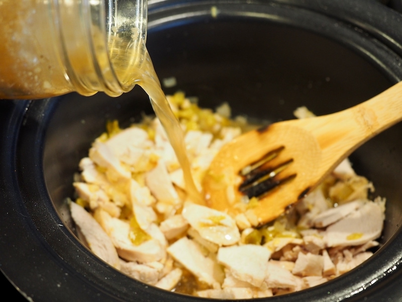pouring in chicken broth from glass jar into crockpot with wooden spoon. 