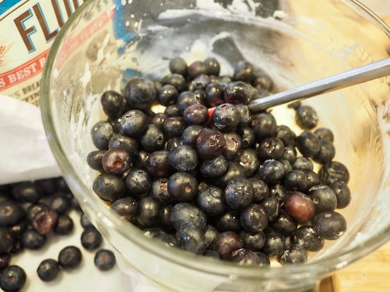Folding fresh blueberries into a glass mixing bowl with blueberries on the counter, red spatula in bowl. 