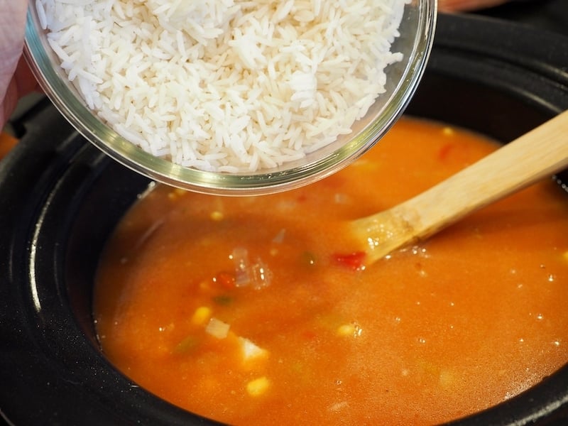 White rice in glass bowl being poured into crockpot of tortilla soup. 