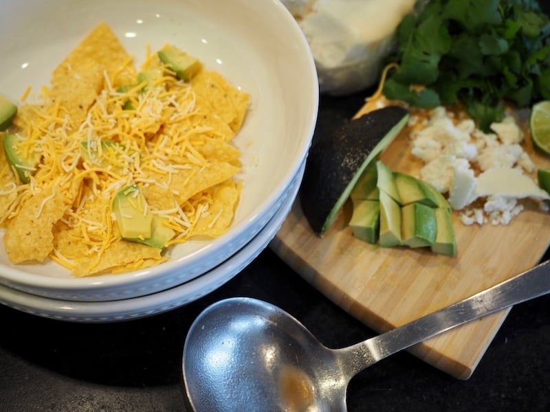 large white bowls with corn tortilla chips, chunks fo avocado and a sprinkle of shredded cheese. Ladle , cheese avocado, lime and cilantro in background.