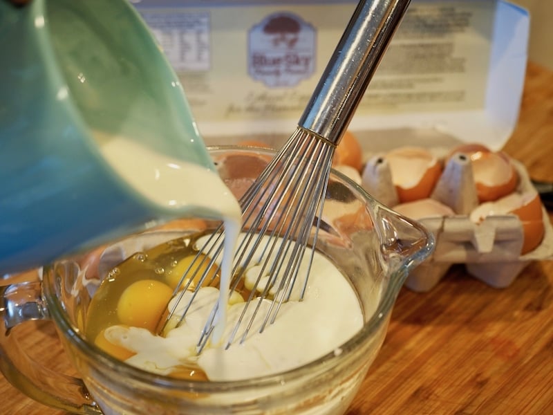 Pouring cream into egg mixture in glass mixing bowl for Prosciutto & Gruyere Quiche with Hash Brown Crust with carton of egg shells in background. 