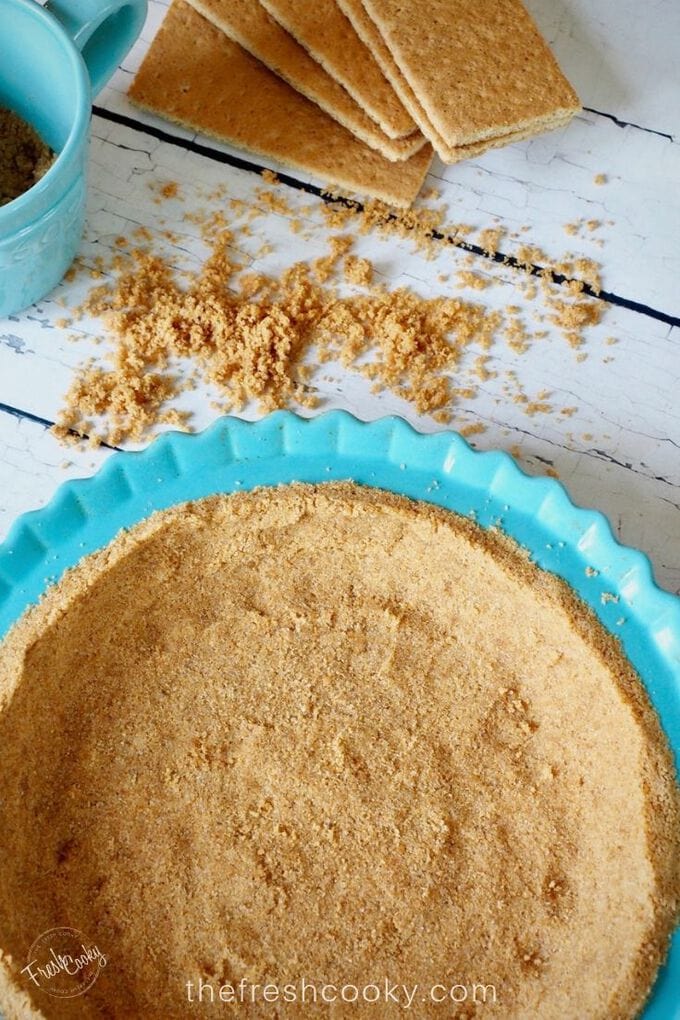 Image of bright turquoise pie plate filled with a graham cracker crust, ready for baking, with crumbs on board behind