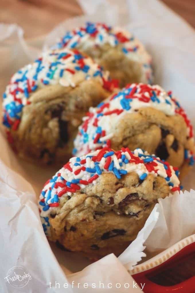 Four Ice Cream Sandwiches using chocolate chip cookies with vanilla ice cream sandwiched between, with red, white and blue jimmies rolled into the ice cream. Laying in a loaf pan lined with white parchment paper. Recipe thefreshcooky.com