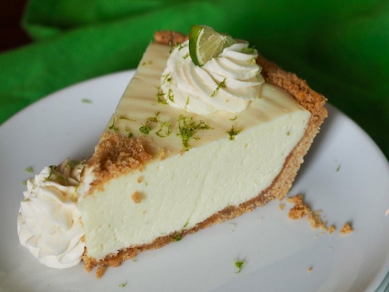 Wedge of key lime pie, topped with creamy whipped cream, lime zest and some extra graham cracker crumbs sitting on a white plate. 