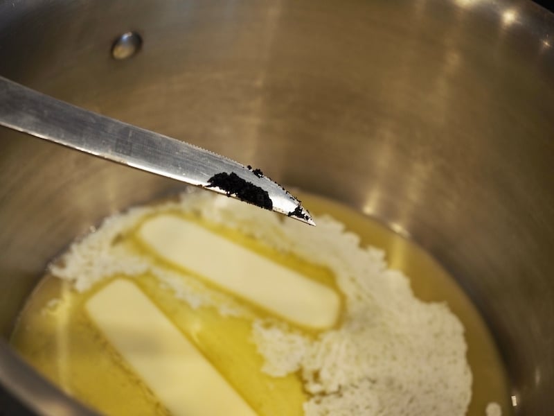A sharp knife with vanilla bean scrapings on it over a pan of melting butter in saucepan. 