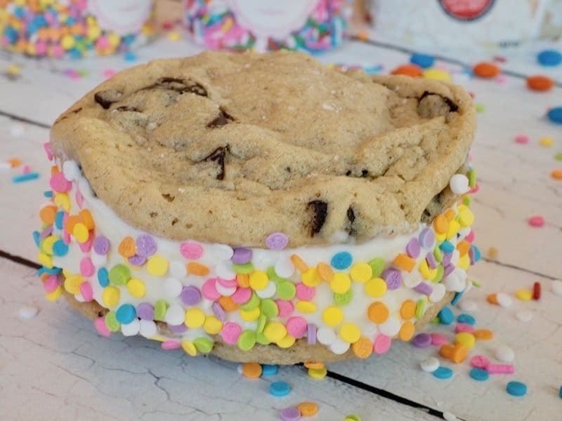 Chewy Chocolate chips cookies with ice cream sandwiched between, rolled in rainbow sprinkles with rainbow sprinkles strewn about the wood. Recipe on thefreshcooky.com