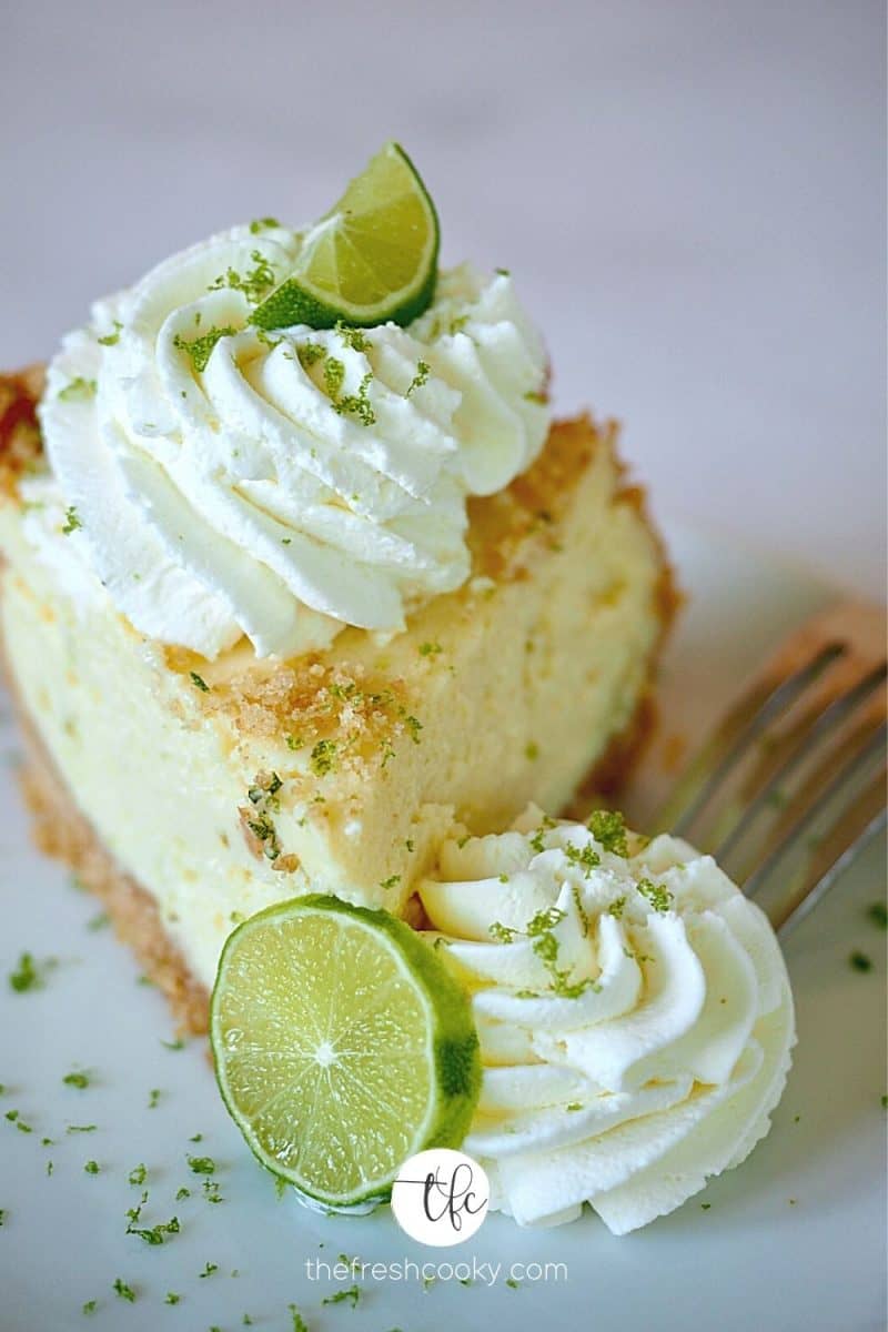 Best Key Lime Pie wedge on a plate with two swirls of whipped cream, garnished with a sliced lime and lime zest.