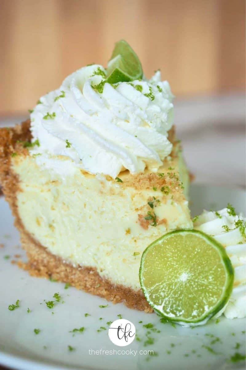 Slice of creamy key lime pie, on white place with two swirls of whipped cream, slice of lime and lime zest sprinkled on top.