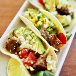 Square image of Mexican Barbacoa Beef street tacos on a white platter with avocado, tomato, cheese and lime.