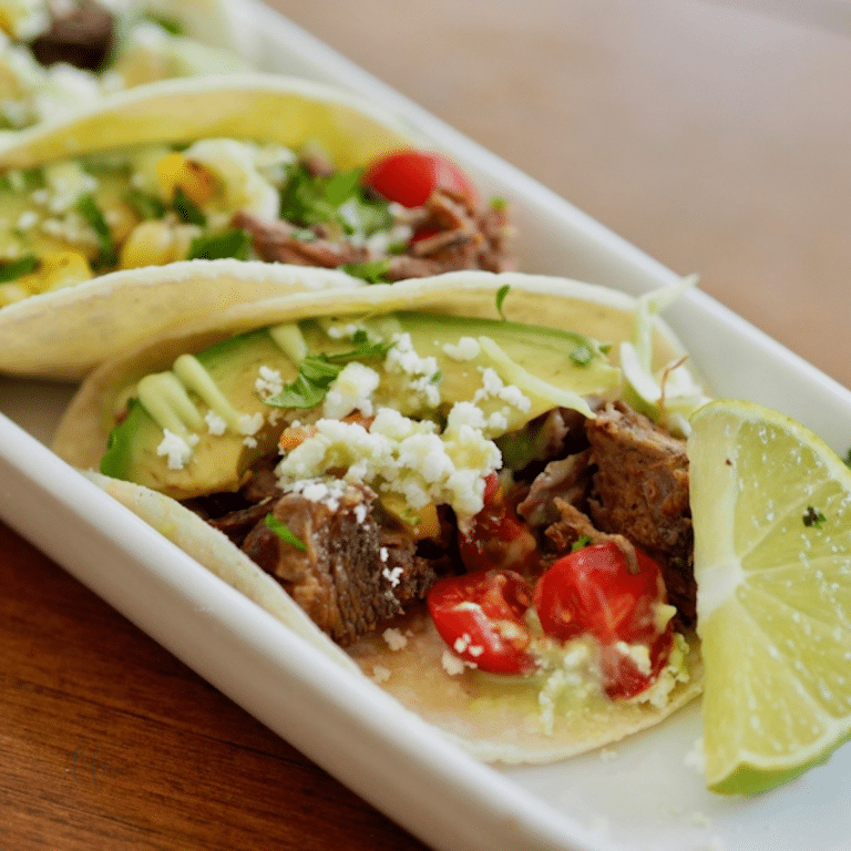 Square image of Mexican Barbacoa Beef in form of street tacos sitting on a white plate with cheese, avocado, tomatoes and lime.
