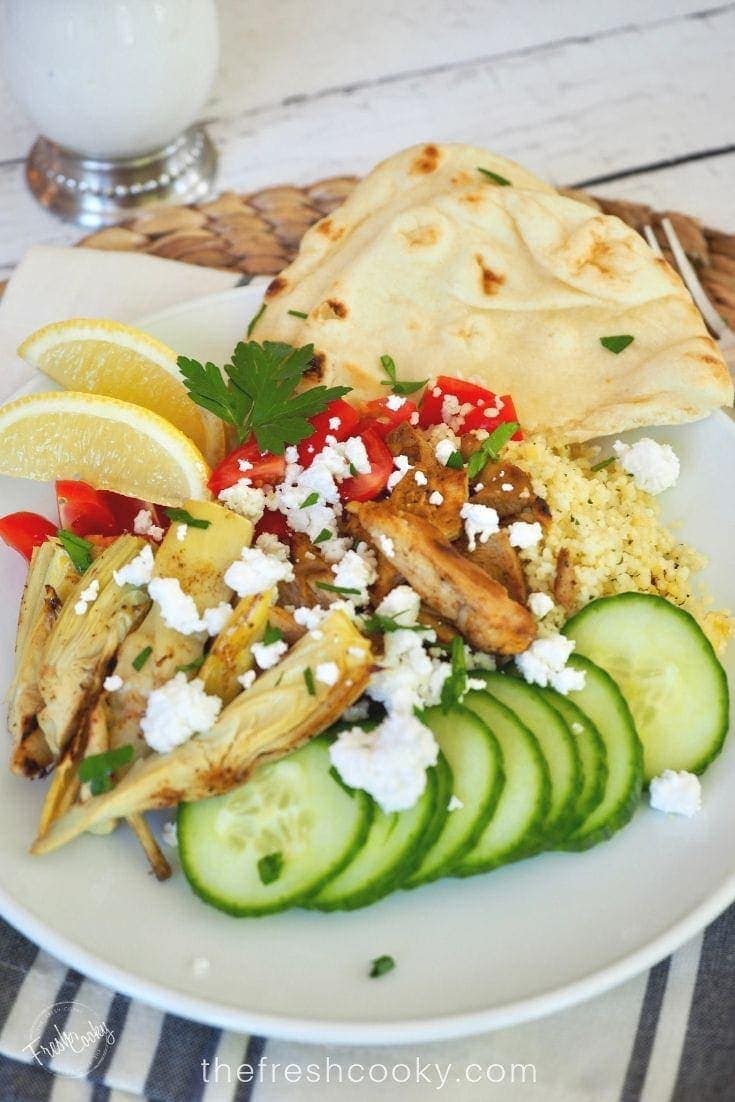 plate filled with sliced chicken shawarma on a bed of cous cous with fresh tomatoes, cucumbers, feta cheese and grilled artichoke hearts, with some naan bread and slices of lemon.