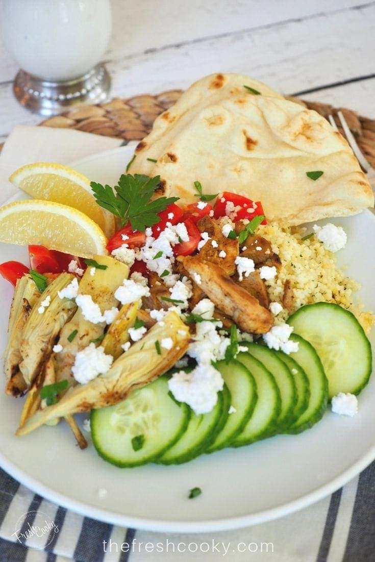 plate filled with sliced chicken shawarma on a bed of cous cous with fresh tomatoes, cucumbers, feta cheese and grilled artichoke hearts, with some naan bread and slices of lemon. 