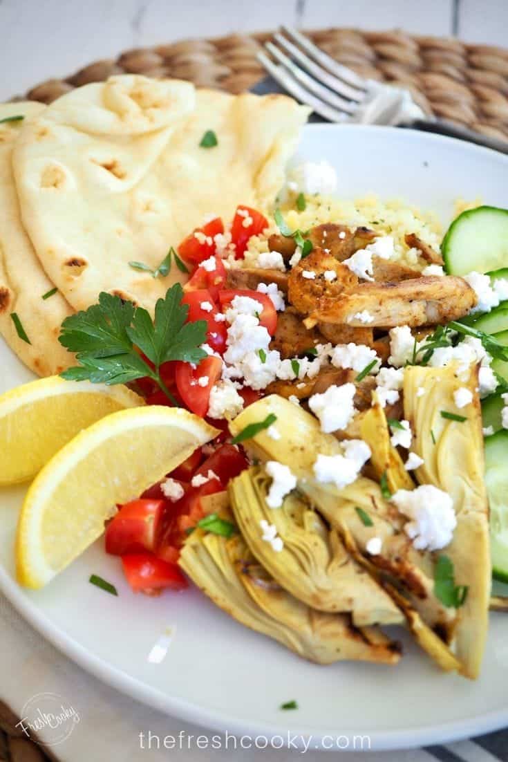 Plate filled with grilled chicken shawarma with artichoke hearts, cucumbers, fresh tomatoes, feta cheese and fresh naan.
