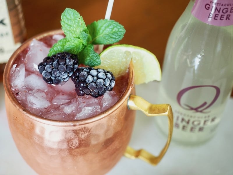 Image of blackberry mule in a copper mug garnished with lime, mint and frozen blackberries, ginger beer bottle in background. 