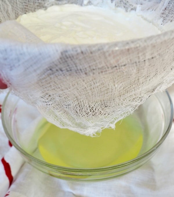 strainer lined with cheesecloth, filled with yogurt, whey collecting in glass bowl below. 
