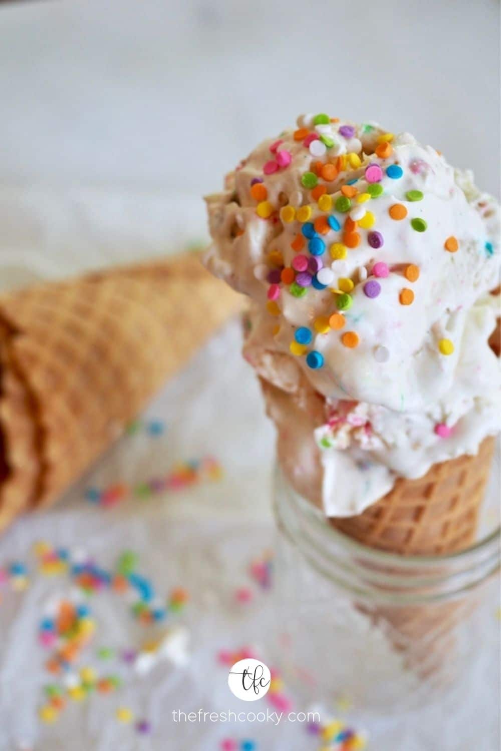 Circus Animal No Churn Ice cream with two scoops on a sugar cone sitting in a glass with sprinkles on top.