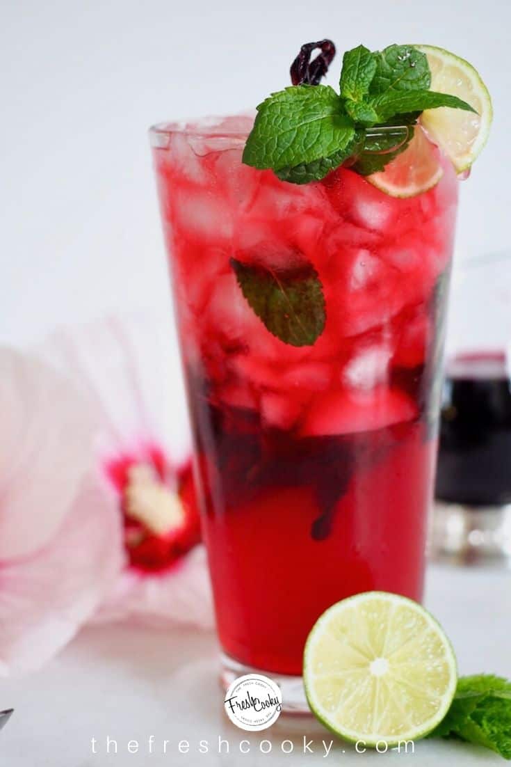 tall glass filled with hibiscus mojito topped with mint, lime slice in foreground and hibiscus flower in background