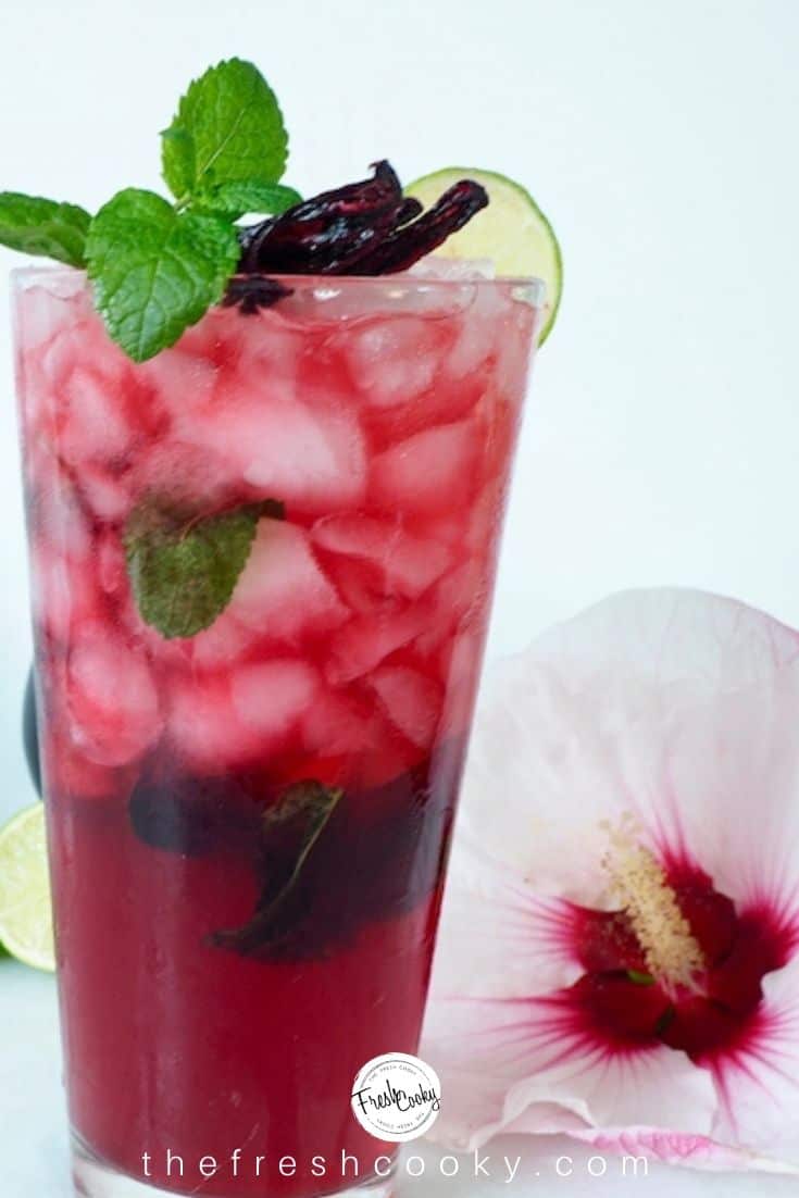 tall glass filled with hibiscus mojito topped with mint, lime slice in foreground and hibiscus flower in background