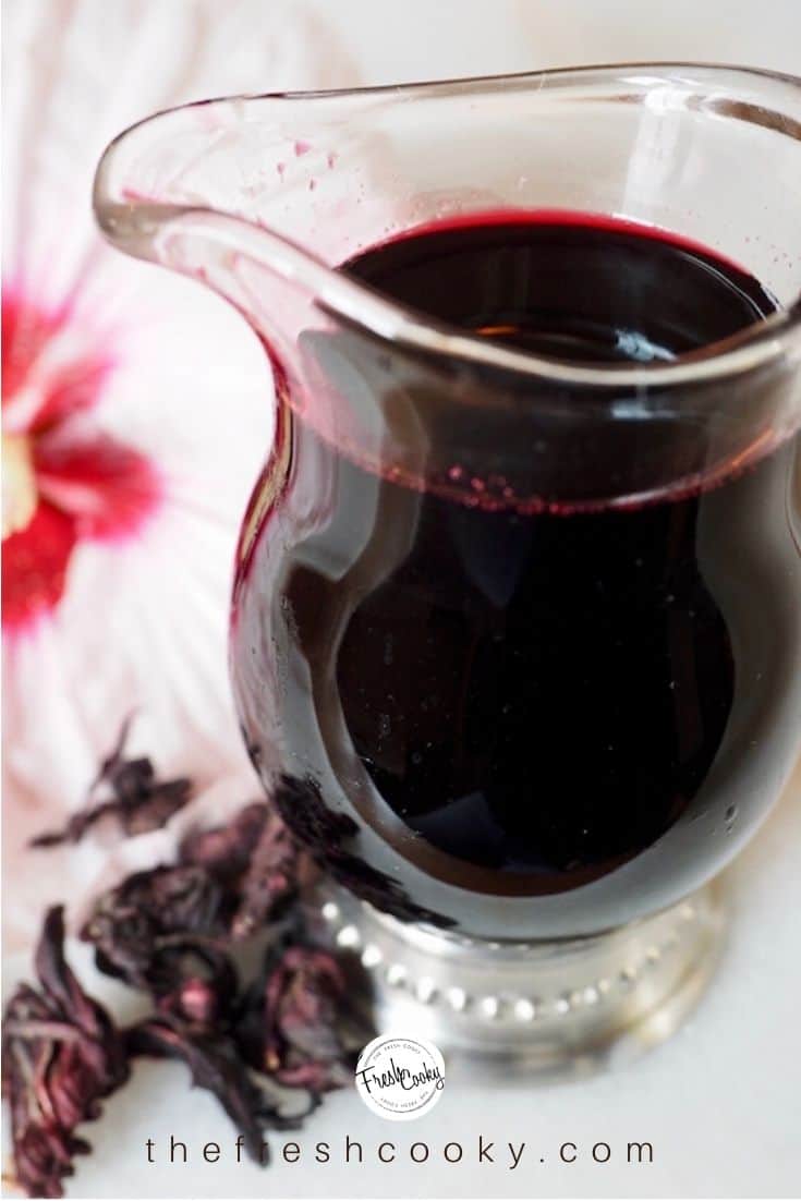 Pin with glass jar of hibiscus simple syrup with dried hibiscus flowers at base and hibiscus flower in background.