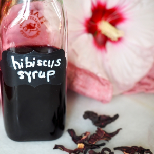 hibiscus syrup recipe in jar with label with dried hibiscus flowers and a fresh hibiscus flower behind.