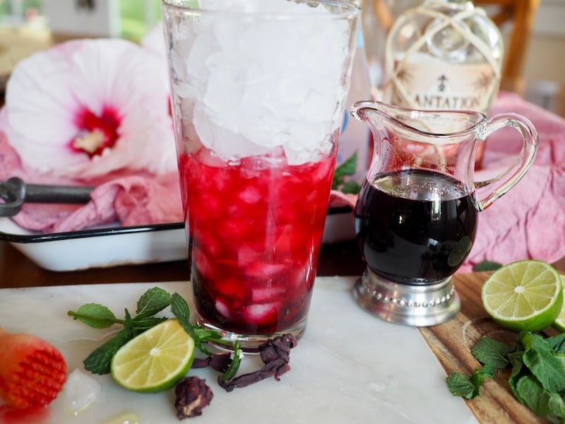 tall glass full of crushed ice, half full with hibiscus simple syrup, rum. Hibiscus syrup in small pitcher, rum bottle and hibiscus flower in background. 