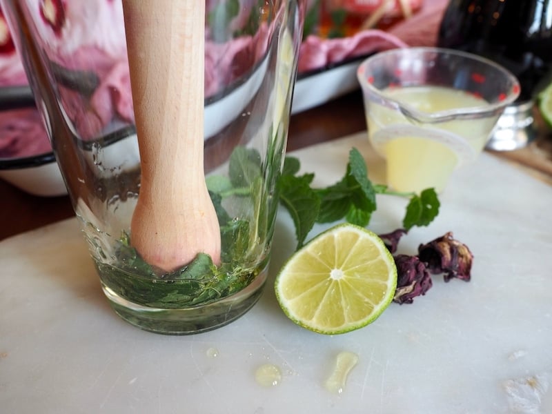 tall glass with mint in bottom and muddler, with agave and slice of lime and lime juice in background.