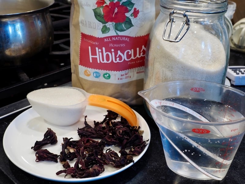 package of dried Hibiscus flowers, canister of sugar, measuring cup of water, measuring cup of sugar and dried hibiscus flowers on plate. 