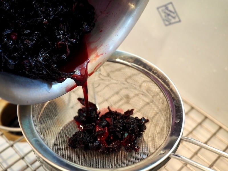 Pouring hibiscus colored water through sieve, catching dried flowers. 