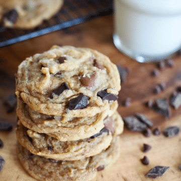 Square Image of triple chocolate chip vanilla pudding cookies on wood table with chocolate chips laying around and a glass of milk in background.
