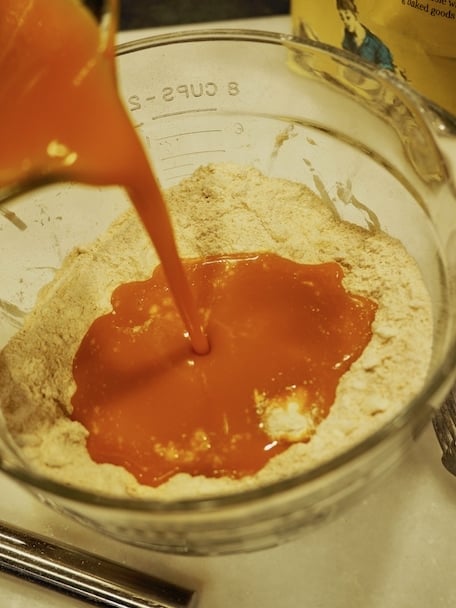 Pouring pumpkin wet mixture into dry flour and sugar mixture in glass mixing bowl on countertop.