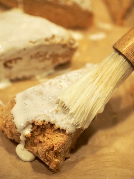 Pastry brush with cream brushing onto uncooked scones on baking sheet.