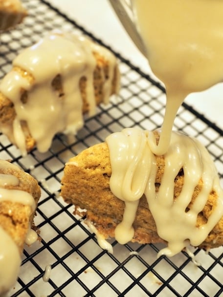 brown butter glaze being drizzled onto pumpkin scones sitting on cooling rack on countertop. 