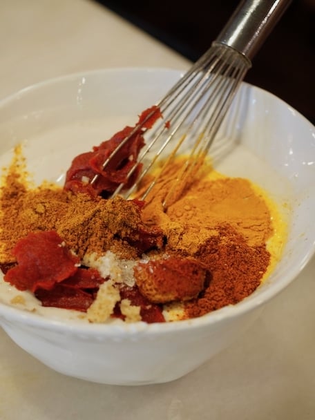 Large white mixing bowl with coconut milk, tomato paste and numerous Indian Spices with a whisk. 