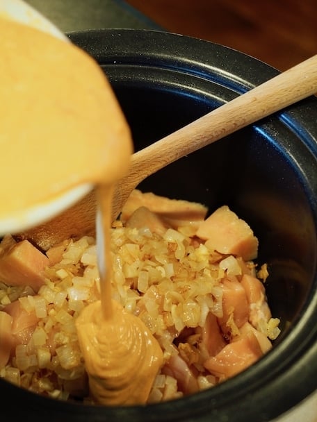 Pouring curry sauce over diced raw chicken and sauted onions and garlic into slow cooker. 