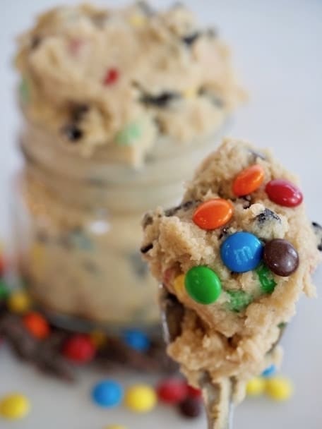 Spoonful of edible cookie dough with 5 colorful M&M's stuck to dough on spoon. Jar of cookie dough overflowing in background with m&m's sprinkled about. 