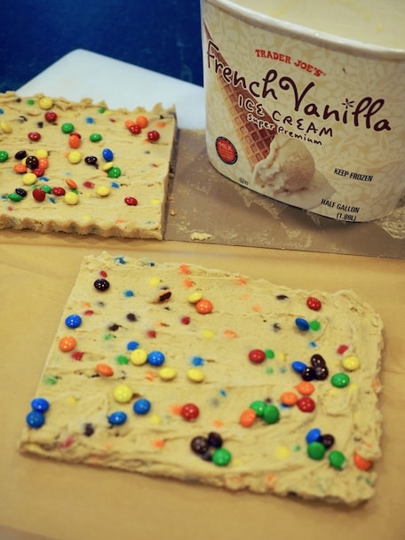 Two "slabs" of frozen cookie dough (safe to eat) with m & m's sprinkled on top cut in half, vanilla ice cream tub in background. 