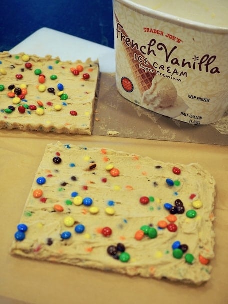 Two "slabs" of frozen cookie dough (safe to eat) with m & m's sprinkled on top cut in half, vanilla ice cream tub in background.